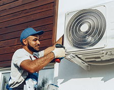 A man wearing blue coveralls and a blue hat stands on a ladder to install a fan.